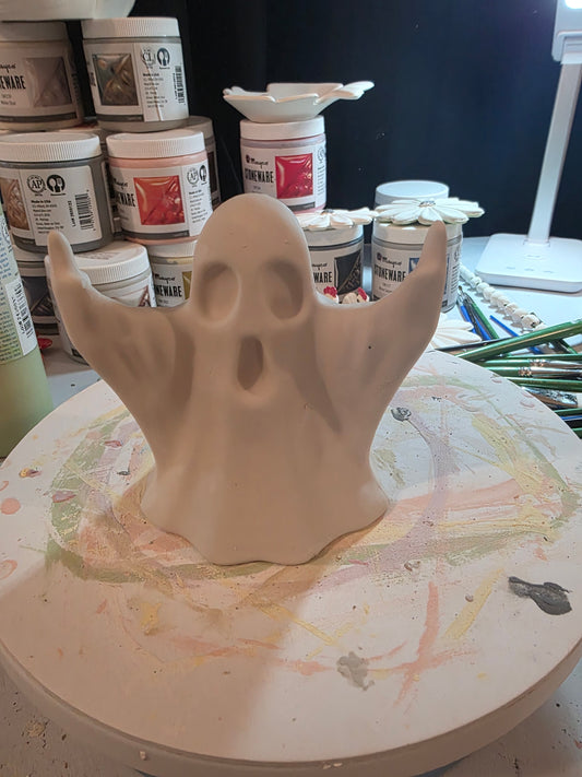 Unpainted ceramic ghost with raised arms on a paint-splattered turntable, surrounded by glaze jars and brushes