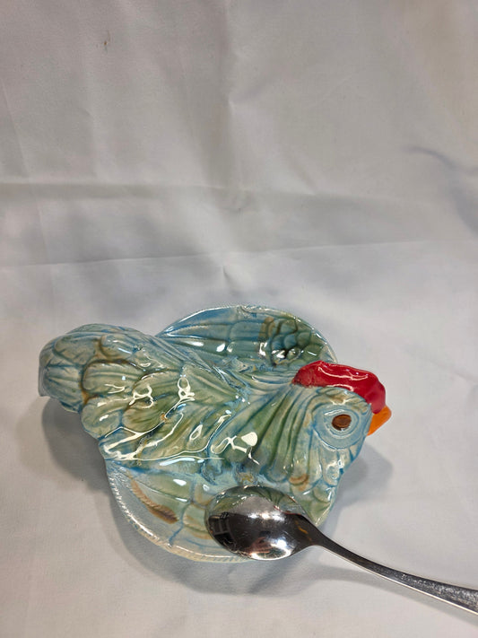 Glossy blue-green ceramic hen spoon rest with a red comb and a metal spoon resting in its curved dish.
