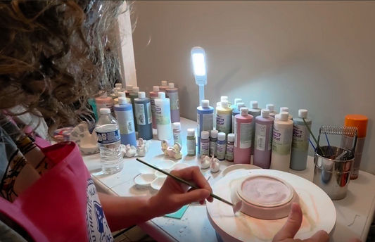 A close-up view of ceramic artist Pottery Pirate sitting at a brightly lit studio desk, carefully painting a ceramic piece on a banding wheel with a fine paintbrush surrounded by bottles of Mayco Stroke & Coat glazes and miniature clay figures.