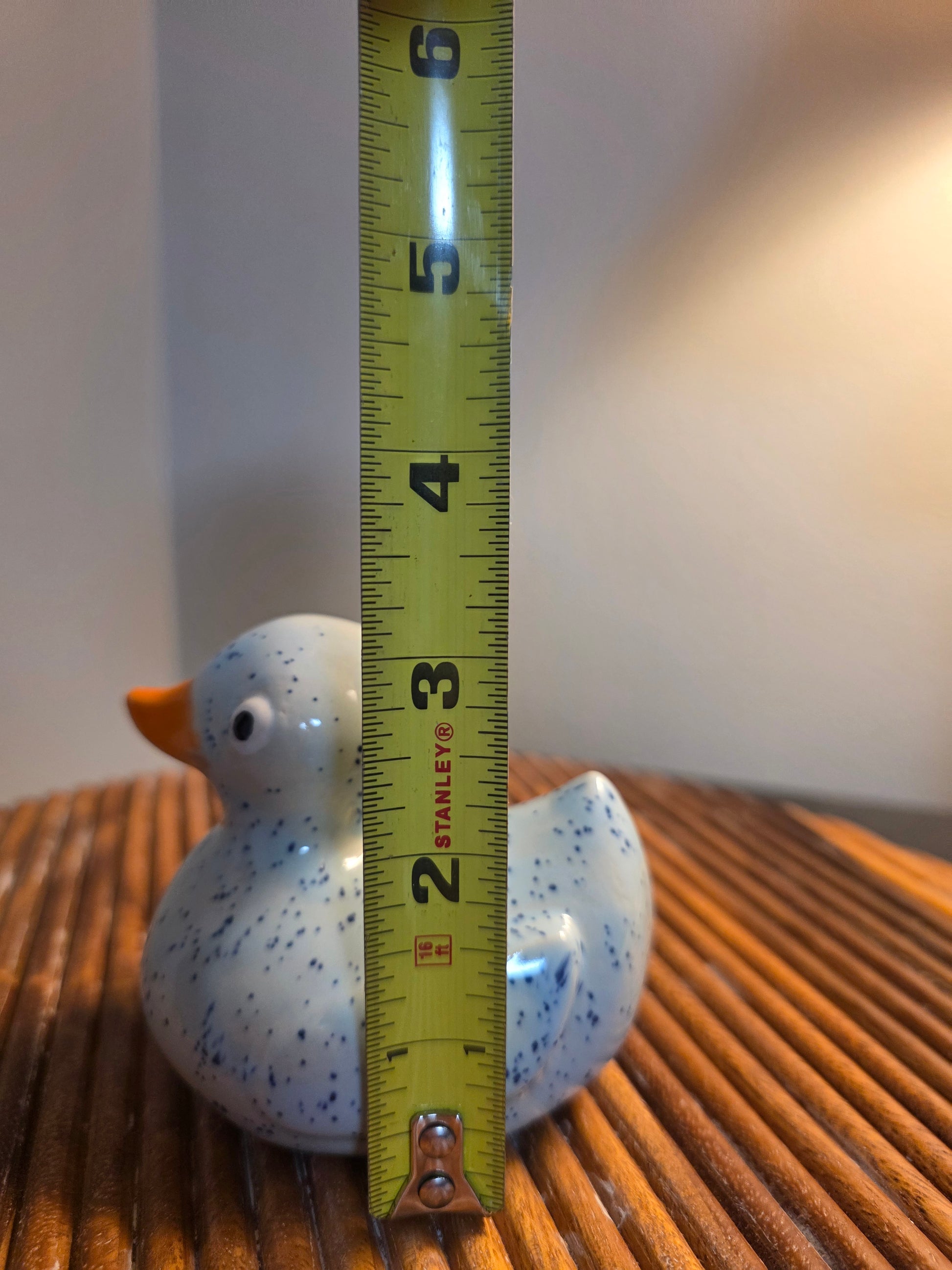 A small speckled white rubber duck with an orange beak standing on a wooden slatted surface, with a yellow Stanley tape measure placed vertically in front of it to show height.