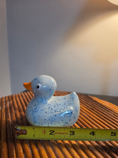 A small ceramic rubber duck figurine in light blue with dark blue speckles and an orange beak, shown next to a yellow measuring tape on a brown slatted surface.