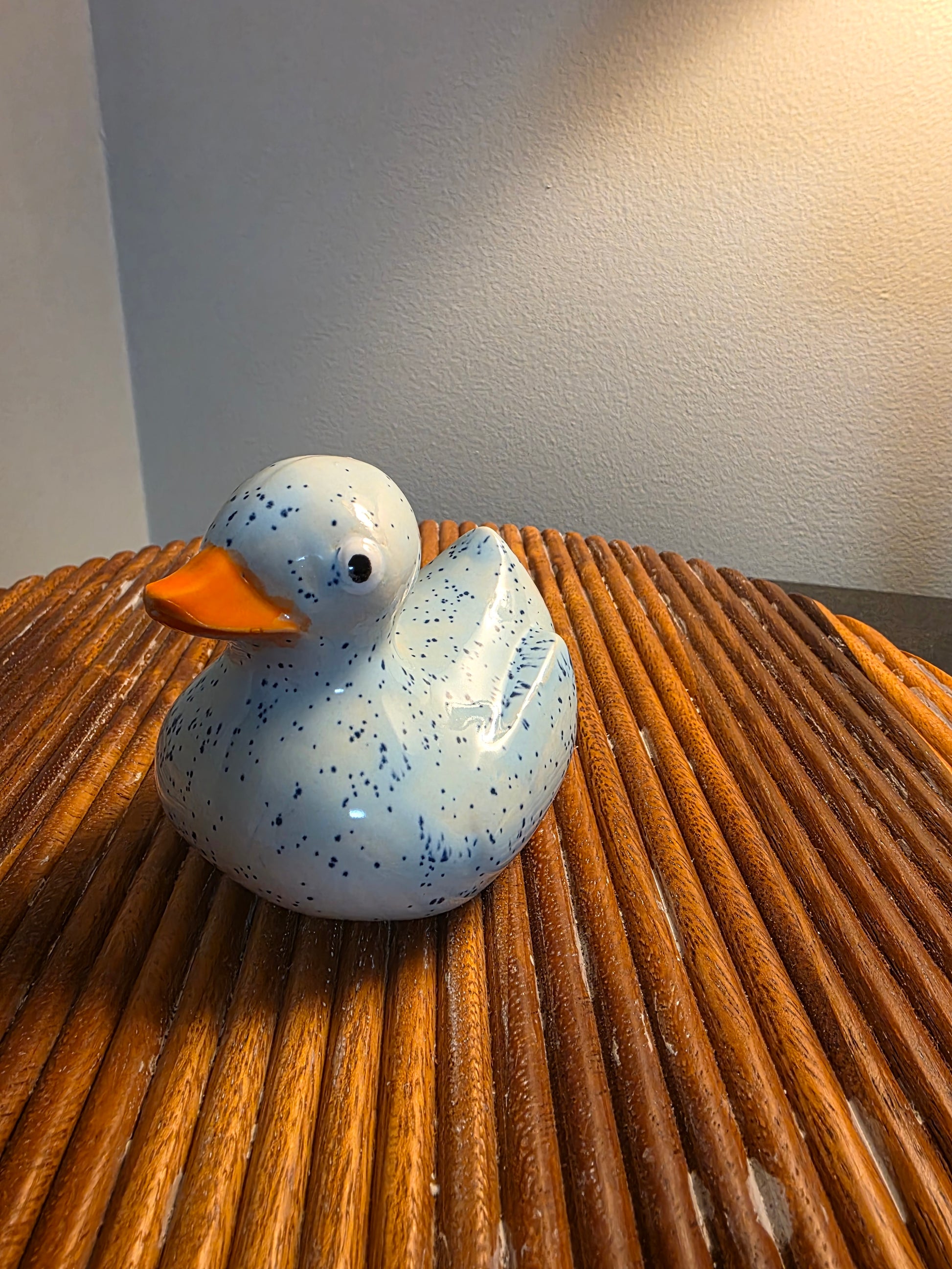 A ceramic duck figurine with a speckled light blue and white glaze and an orange beak, sitting on a round wooden slatted surface against a neutral wall.