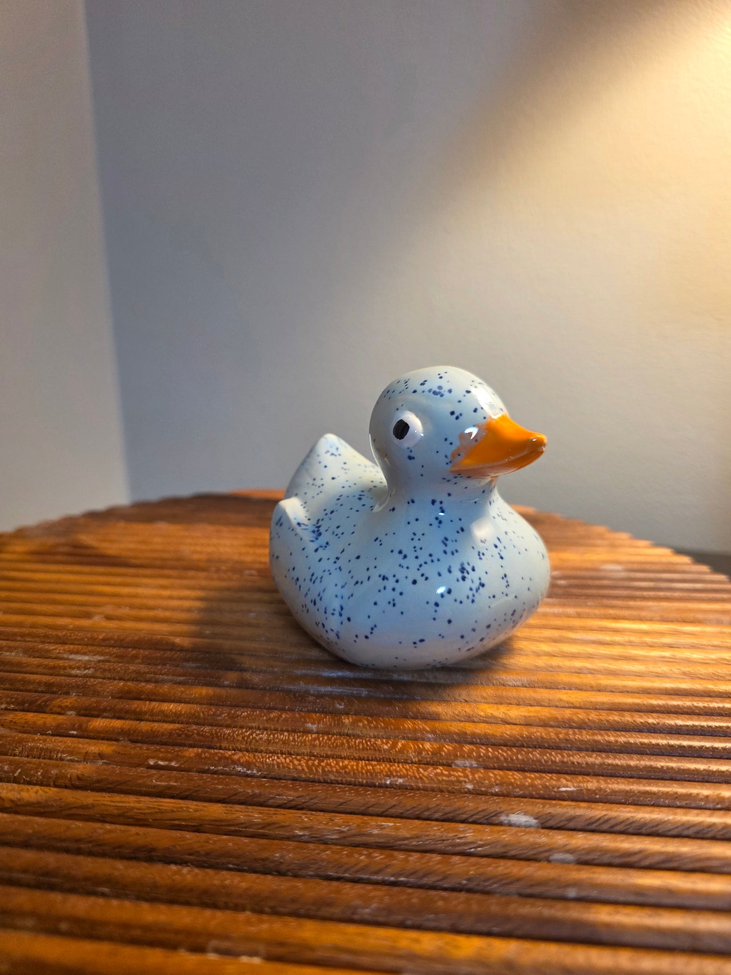 A small speckled rubber duck toy in light blue with a bright orange beak, sitting on a brown slatted wooden surface against a neutral background.