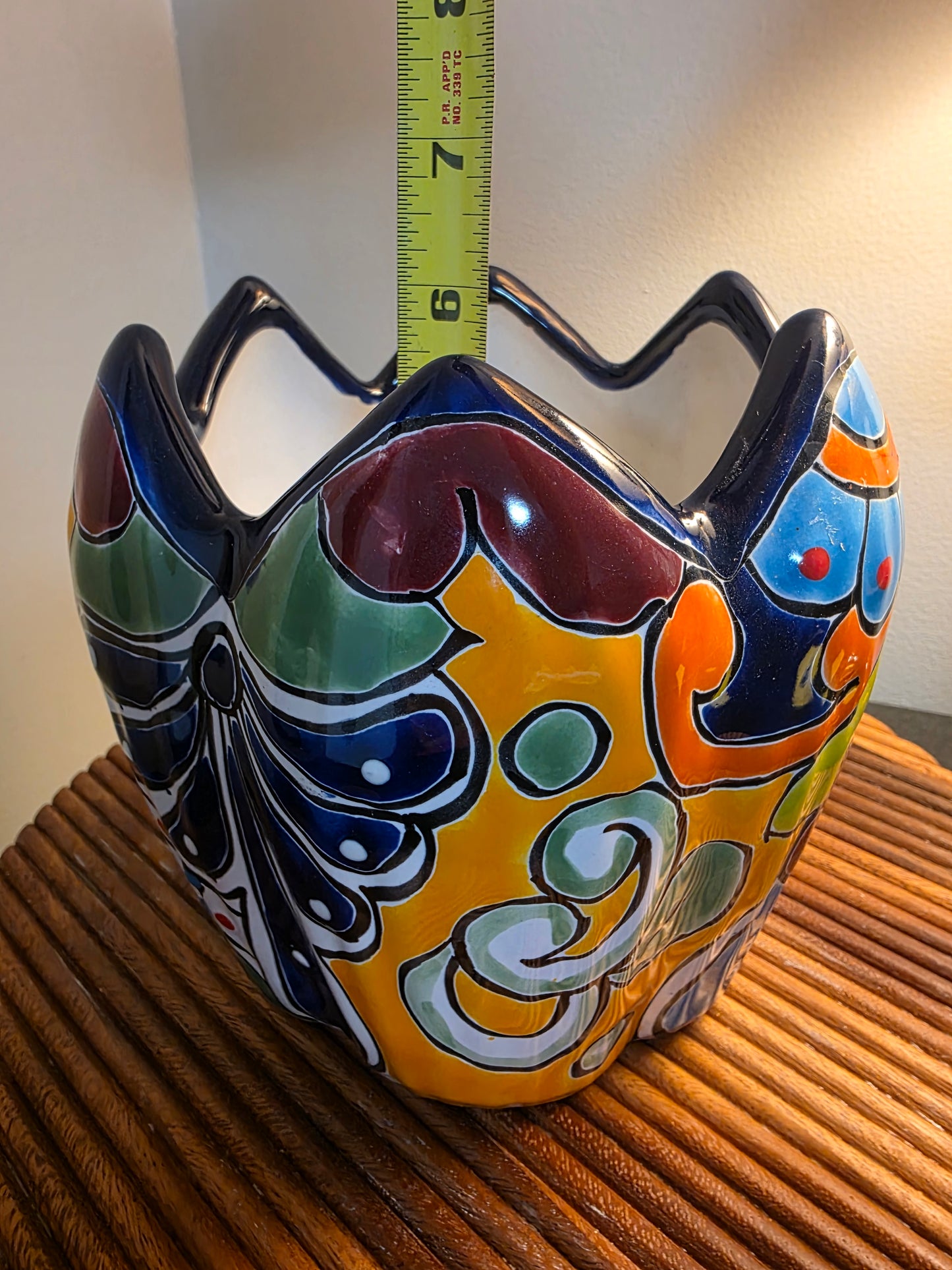 A colorful hand-painted ceramic planter with a yellow, blue, green, and orange pattern, shown next to a yellow tape measure and a brown ribbed surface.