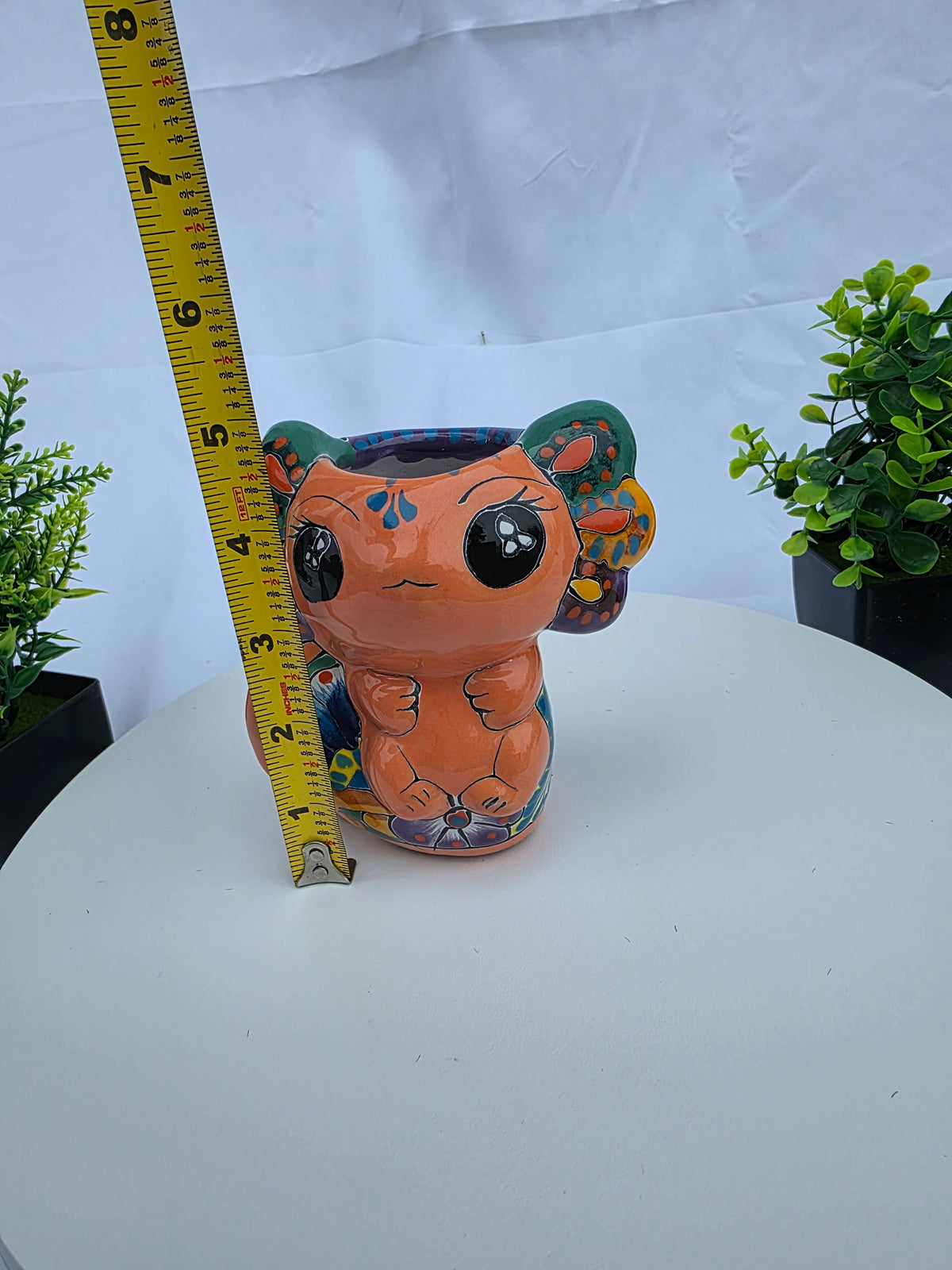 A small hand-painted ceramic axolotl figurine in terracotta orange with colorful floral patterns, shown next to a yellow tape measure on a white surface with a white backdrop and small potted plants on either side.