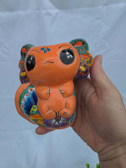 A hand holding a small, hand-painted ceramic AXOLOTL figurine with a bright orange body and colorful, patterned details on its ears, back, and paws, set against a wrinkled white fabric background.
