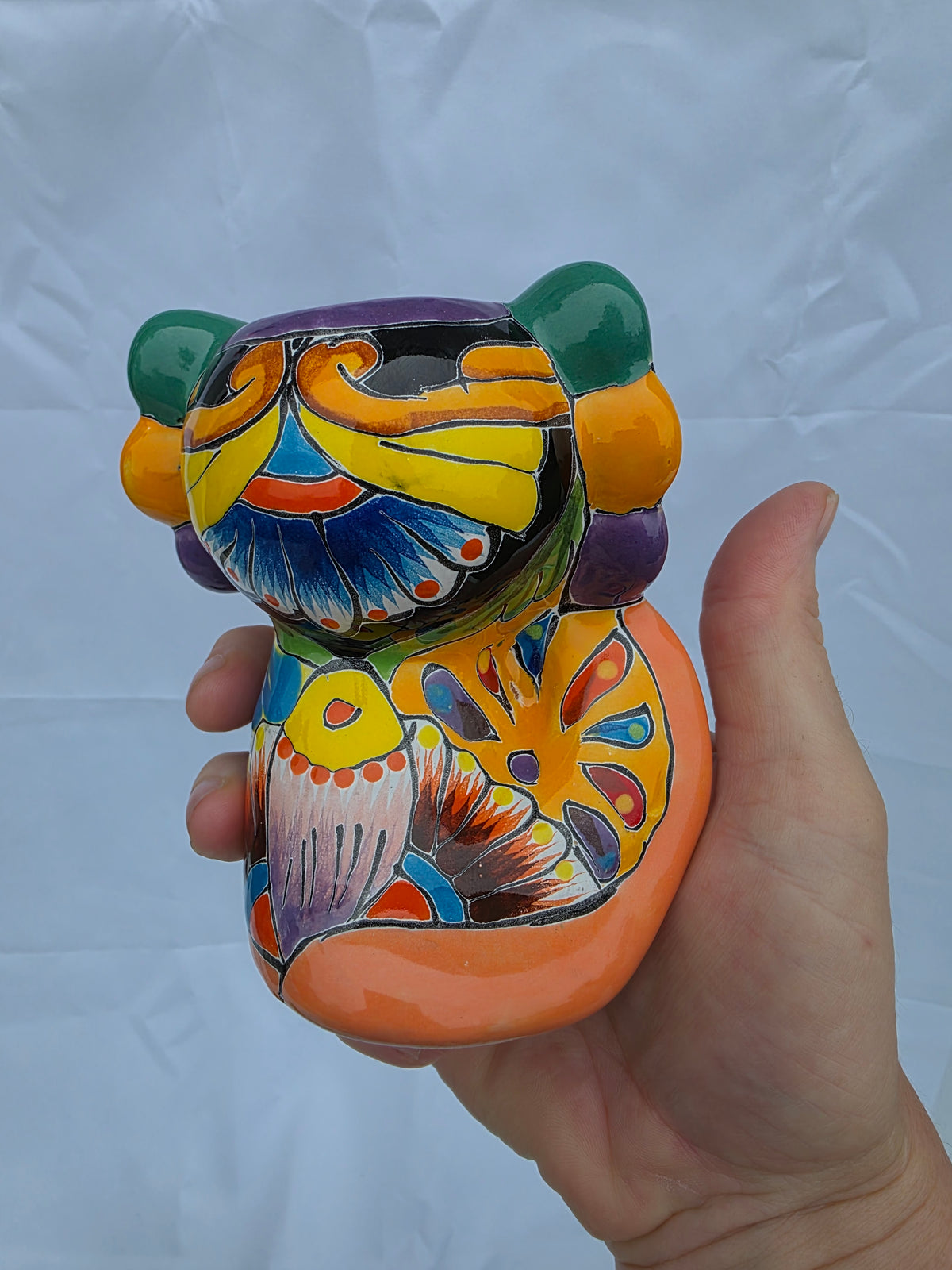 A hand holding a small hand-painted ceramic axolotl figurine decorated with vibrant traditional Mexican-style patterns in orange, yellow, blue, green, purple, and white against a light gray wrinkled backdrop.