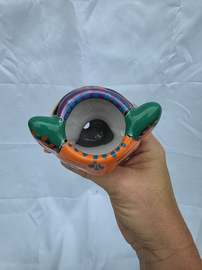 A hand holding a colorful ceramic axolotl planter with green handles, an orange base, and blue and purple decorative patterns, set against a wrinkled white fabric background.