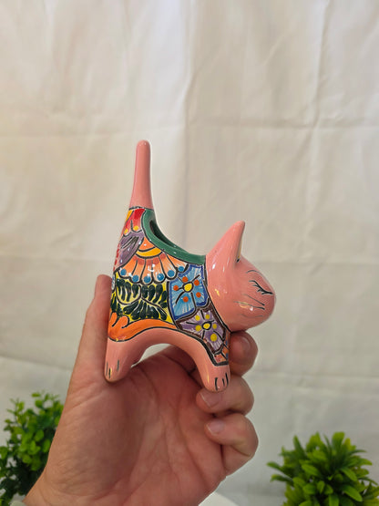 Hand holding a colorful ceramic cat figurine on a white surface with green plants in the background.