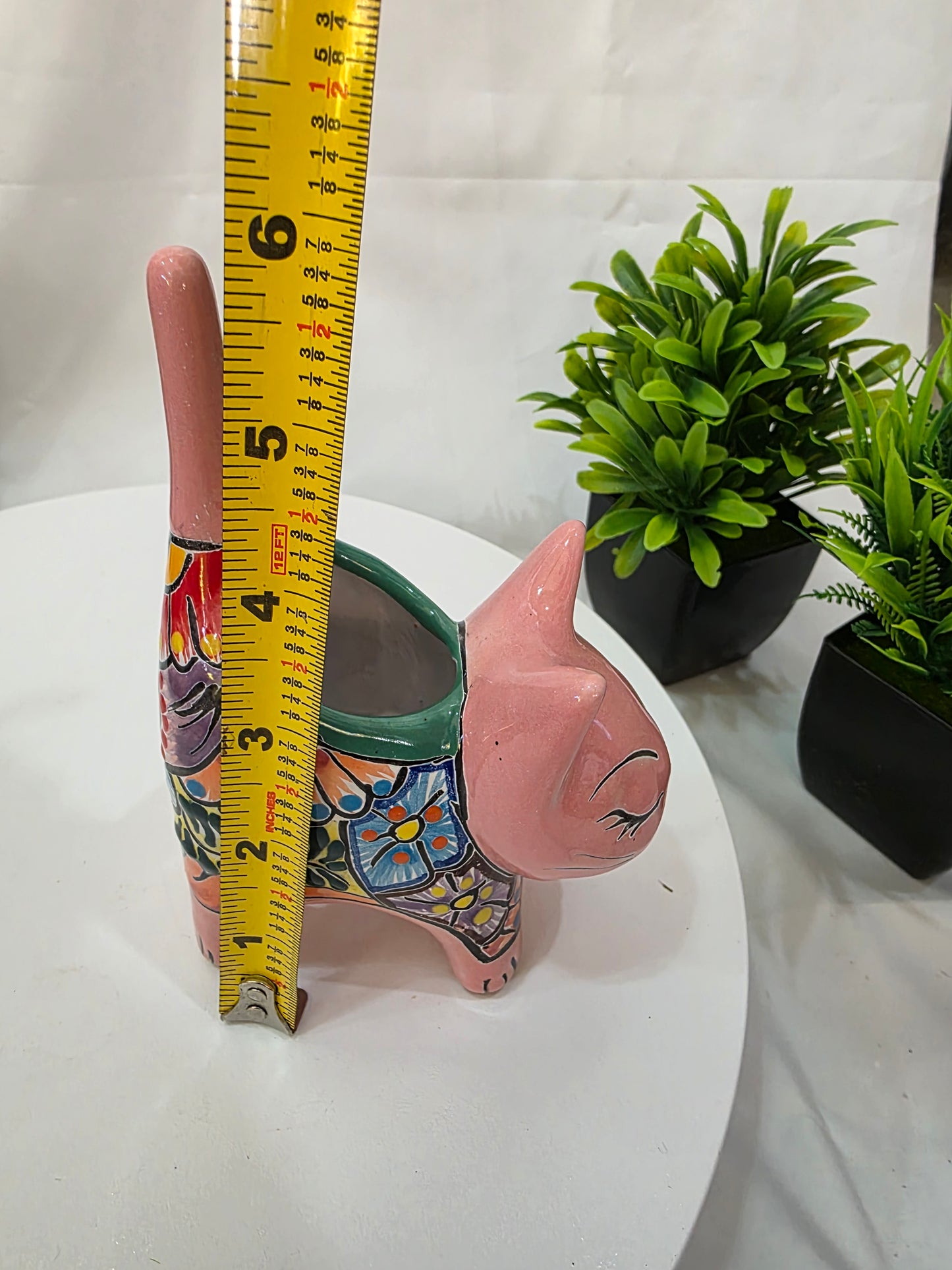 Decorative pink cat planter with a plant on a white surface, measured by a yellow tape measure.