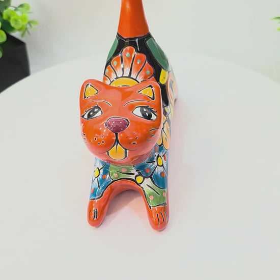 A 360-degree rotating view of a handcrafted Mexican Talavera ceramic cat figurine lying down on a white surface. The cat is painted in a vibrant orange with a glossy finish, featuring detailed multi-colored floral and geometric folk art patterns in yellow, blue, and green across its back. Its long, straight orange tail serves as a functional ring holder
