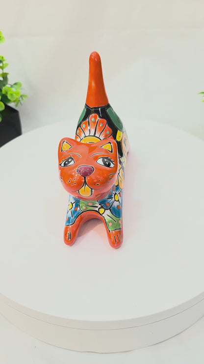 A 360-degree rotating view of a handcrafted Mexican Talavera ceramic cat figurine lying down on a white surface. The cat is painted in a vibrant orange with a glossy finish, featuring detailed multi-colored floral and geometric folk art patterns in yellow, blue, and green across its back. Its long, straight orange tail serves as a functional ring holder
