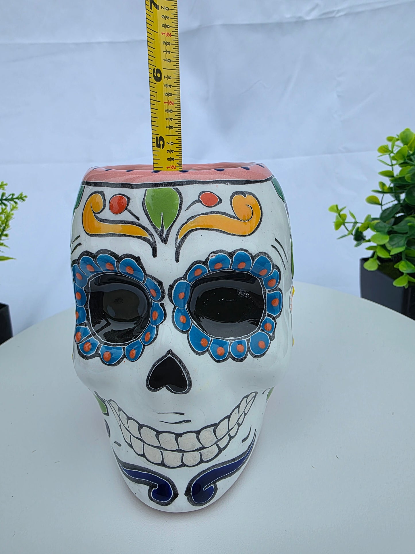 A hand-painted ceramic sugar skull with Day of the Dead (Dia de los Muertos) style designs, shown with a yellow tape measure on a white background.