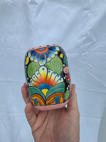 A hand holding a small cylindrical ceramic cup decorated with a colorful hand-painted folk art pattern on a wrinkled white fabric background.