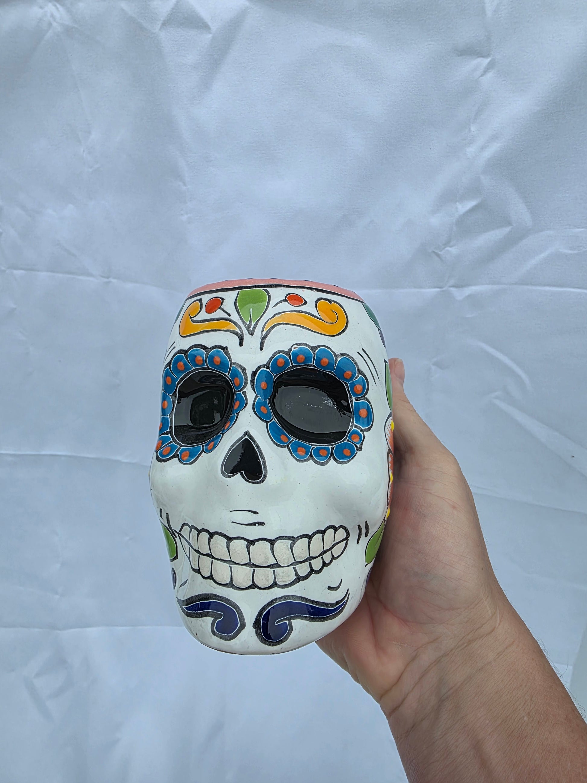 A hand holding a white ceramic sugar skull container decorated with colorful Day of the Dead patterns, set against a wrinkled white fabric background.