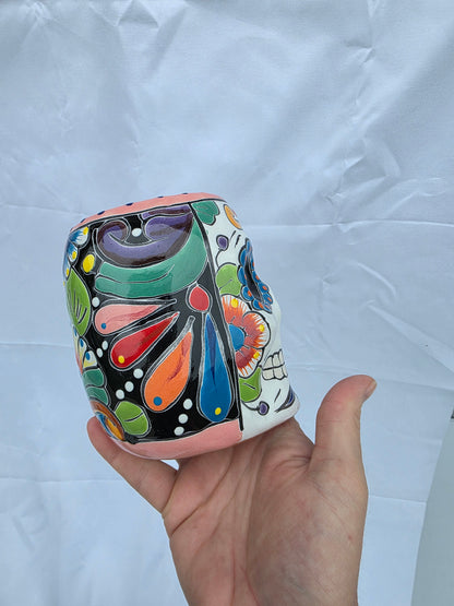 A hand holding a small cylindrical ceramic cup decorated with a colorful hand-painted folk art pattern on a wrinkled white paper background.