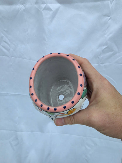 A hand holding a ceramic planter pot with a pink rim decorated with dark blue polka dots, shown from a top-down view against a wrinkled white background.